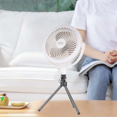 A small white personal fan on a mini tripod stand, shown in a bright living room setting with a person sitting on a white sofa in the background.