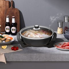 A black electric hot pot with a clear glass lid sits on a gray countertop, filled with a simmering chicken broth. The scene is styled as a kitchen or dining setup with various food props, including plates of food, bottles of beer, wooden cutting boards, and spice grinders.